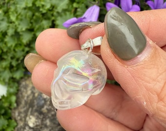May include: A clear, iridescent skull pendant with a silver bail. The skull is carved with detailed features and catches the light, displaying a rainbow effect. The pendant is held against a backdrop of green foliage and purple flowers.