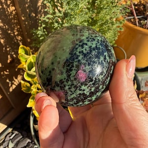 Puede incluir: Una esfera pulida de zoisita verde con inclusiones negras y rubíes. La gema se sostiene en una mano, con un fondo borroso de vegetación y una maceta de terracota. La superficie de la esfera refleja la luz, mostrando sus patrones y colores naturales.