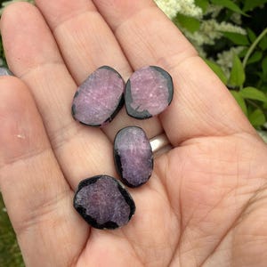Puede incluir: Tres pequeñas rodajas de piedras preciosas pulidas, planas y de formas irregulares y redondeadas. Cada pieza presenta un centro rosa translúcido y texturizado rodeado por un borde exterior negro opaco, mostrando un aspecto mineral bicolor.