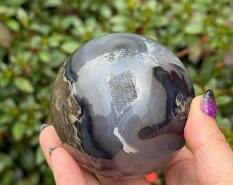 Peut inclure: Une sphère en agate polie avec des bandes grises, noires et blanches. La boule de cristal est tenue dans une main, mettant en valeur sa surface lisse et réfléchissante. La sphère présente une cavité semblable à une géode. Le fond est un feuillage vert flou.