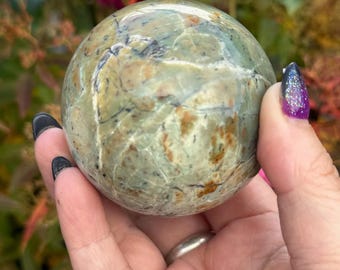 May include: A green sphere with brown and black veining. The polished stone is held in a hand with a silver ring and dark nail polish. The background features out-of-focus pink and green foliage.