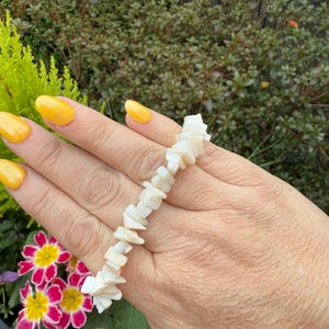 Op de afbeelding: Een witte schelpenarmband gemaakt van kleine, onregelmatige schelpenstukken. De armband zit om een pols.