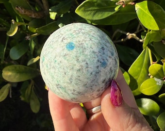 May include: A round, speckled sphere with a light green and blue color scheme. The sphere is held in a hand with a painted fingernail. The background features green leaves and sunlight.