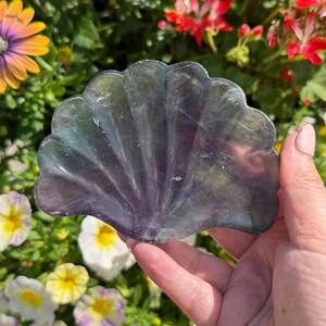 May include: A hand holds a translucent, shell-shaped object with a scalloped edge. The object has a gradient of colours, including purple, green, and clear. The background features colourful flowers, including yellow, white, and red blooms.