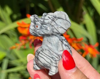 May include: A small, grey elephant figurine with black veining. The elephant is carved from stone and has a trunk, ears, and legs. The background features green foliage and orange flowers.