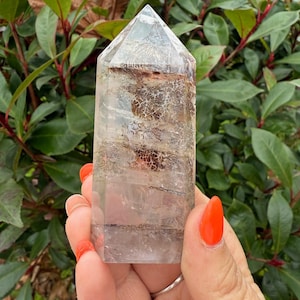 Könnte beinhalten: Eine klare Quarzkristallspitze mit braunen und weißen Einschlüssen. Der Kristall ist oben spitz zulaufend und hat eine flache Basis. Er ist etwa 12,5 cm hoch.