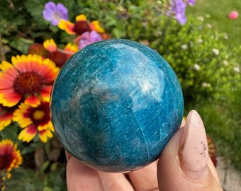 May include: A close-up of a teal-colored crystal sphere. The sphere is held in a hand, with a blurred background of colorful flowers and greenery. The sphere has a smooth, polished surface.