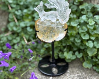May include: A clear carved crystal fox with a large, detailed tail sits atop a rounded, golden-yellow crystal base. The sculpture is supported by a black metal stand with a circular base. A circular logo in the corner reads CRYSTAL PARADISE.