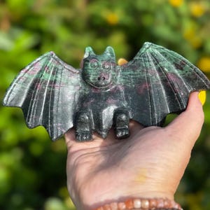 May include: A carved stone bat figurine with outstretched wings. The bat is primarily dark grey with streaks of green and red. The figurine is held in a hand against a blurred green and yellow background.