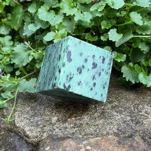 Può includere: Un blocco quadrato color verde acqua con macchie scure, appoggiato su una superficie di pietra. Il blocco ha un aspetto strutturato ed è circondato da fogliame verde. La forma cubica suggerisce un oggetto decorativo o artistico.