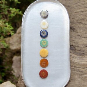Puede incluir: Siete piedras de chakra dispuestas verticalmente en una bandeja ovalada blanca. Las piedras son redondas y presentan símbolos dorados. Las piedras son, de arriba a abajo, transparentes, blancas, azules, verdes, amarillas, naranjas y rojas.