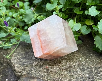 May include: A faceted, translucent crystal cube with peach and white hues and reddish-orange internal striations. The crystal is resting on a mossy, textured rock surface, with green foliage in the background. This natural crystal is a decorative item.
