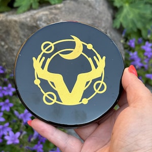 May include: A black round coaster with a gold celestial design featuring a crescent moon, hands, and circles. The coaster is held in a hand with red nail polish, with purple flowers in the background.