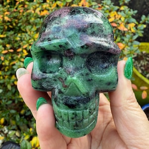May include: A carved skull made from green and maroon stone. The skull features detailed eye sockets, a nose, and a jawline with teeth. The stone has a polished finish, and the skull is held in a hand.