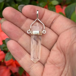 May include: A clear quartz crystal pendant with a silver-colored metal cap and bail. The crystal is a long, hexagonal shape. The pendant is held in a hand against a background of green foliage and red flowers.