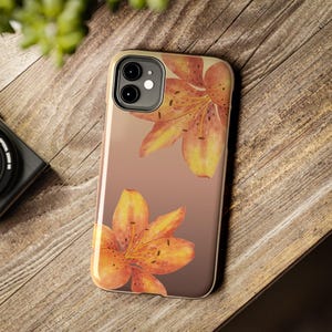 Puede incluir: Una funda para smartphone con un fondo degradado suave que transita de beige claro a marrón oscuro, decorada con dos grandes flores de lirio de color naranja vibrante con manchas marrones oscuras en las esquinas superior derecha e inferior izquierda.