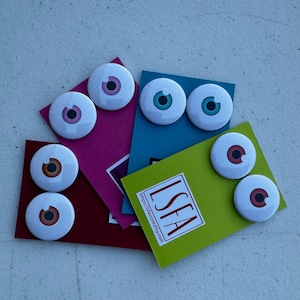 May include: Six white round buttons with different colored eyes printed on them. The buttons are arranged on four different colored cardstock backgrounds. The cardstock is folded in half and the buttons are attached to the front of the cardstock. The cardstock is green, blue, pink, and red. The green cardstock has the text "ISFEA" and a website address printed on it.