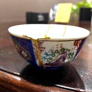 May include: A small ceramic bowl featuring a white interior and a decorative exterior with blue, red, green, and purple floral patterns. The bowl has a visible crack repaired with gold lacquer, highlighting the kintsugi technique on the rim.