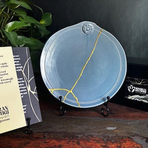 May include: A round, light blue ceramic plate featuring a prominent gold-colored kintsugi repair line running diagonally across the surface. The plate has a small circular decorative emblem at the top and rests on a black metal display stand.