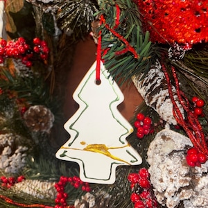 May include: A white ceramic Christmas tree ornament with a green outline and a gold painted accent across the center. The ornament hangs from a red ribbon against a background of pine branches, frosted pinecones, and red berries.