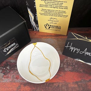 May include: A white ceramic bowl featuring a gold-colored kintsugi repair line, accompanied by a black gift box with a Sonoran Kintsugi logo, a yellow informational card, and a black greeting card with the text Happy Anniversary in white script.