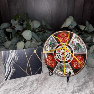 May include: A decorative ceramic plate featuring a segmented pattern of red, black, and white floral and carriage motifs, displayed on a black metal stand. Beside it sits a rectangular card with a black background, gold lines, and white dots.