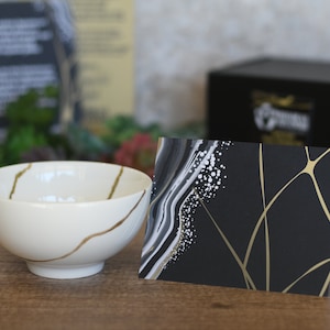 May include: A white ceramic bowl featuring gold-colored kintsugi-style lines and a folded greeting card with a black background, gold geometric lines, and a white and gray marbled design on the left side, placed next to a black gift box.