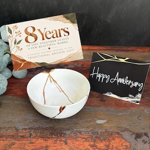 Könnte beinhalten: Eine weiße Keramikschale mit goldfarbenen Rissdetails, begleitet von zwei Karten. Eine Karte trägt die Aufschrift 8 Years of Life Together Leaves a Few Beautiful Marks, die andere Karte zeigt Happy Anniversary in weißer Schreibschrift.