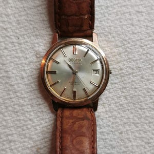 Puede incluir: Reloj de pulsera Dogma vintage con correa de cuero marrón. La esfera plateada tiene agujas e indicadores dorados, y una pantalla de fecha. La correa es de cuero marrón texturizado.