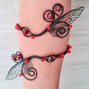 May include: Two black wirework arm cuffs with red beads and stylized wing designs. The cuffs are decorated with red beads and feature intricate wing-shaped details, creating a delicate and whimsical aesthetic.