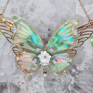 Könnte beinhalten: Eine goldfarbene Schmetterlings-Anhänger-Halskette mit schillernden grünen Flügeln. Die Flügel haben ein zartes, geädertes Design. Eine weiße Blume mit einem Kristallmittelpunkt ziert die Mitte, und grüne Kristallperlen hängen an den Seiten. Die Halskette ist ebenfalls goldfarben.