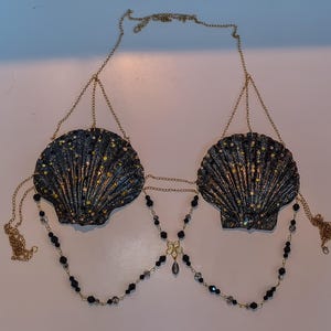 May include: A seashell-shaped top with a gold chain and black bead accents. The shells are dark with glitter and iridescent accents. The top is designed to be worn as a bra.