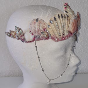 Könnte beinhalten: Eine Krone aus Muscheln mit rosa und weißen Akzenten. Die Krone ist mit Perlen und Glitzer verziert. Eine silberne Kette mit einem Perlenanhänger hängt von der Krone.