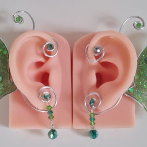 Könnte beinhalten: Ein Paar Feen-Ohrklemmen mit grün glitzernden Flügeln und hängenden grünen Perlen. Die Ohrklemmen sind aus Silberdraht gefertigt und umschließen das Ohr. Die Flügel sind transparent grün mit Glitzer.