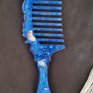Op de afbeelding: Een blauwe harskam met ingebedde glitter en kleine schelpaccenten. De kam heeft een decoratief handvat en een witte kwast. De kam is ongeveer 15 cm lang.