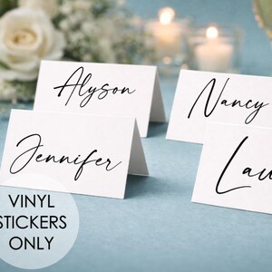 Könnte beinhalten: Weiße Tischkarten mit Namen in eleganter schwarzer Schrift, darunter "Alyson", "Nancy", "Jennifer" und "Laura". Die Karten sind auf einer hellblauen Oberfläche angeordnet, mit dem Text "VINYL STICKERS ONLY" in einem Kreis. Champagnergläser und Blumen sind im Hintergrund.