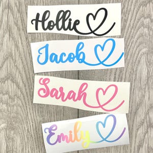 Puede incluir: Cuatro calcomanías con nombres: "Hollie" en negro, "Jacob" en azul, "Sarah" en rosa y "Emily" en colores del arcoíris. Cada nombre va seguido de un diseño en forma de corazón. Las calcomanías están sobre un fondo blanco, con una superficie de madera.