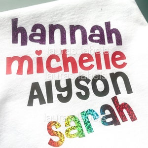 May include: White t-shirt with four names written in glitter: Hannah, Michelle, Alyson, and Sarah. The names are in different colours and styles.
