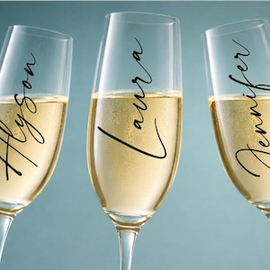 May include: Three champagne flutes filled with sparkling wine. Each glass has a name written in elegant black script: Alyson, Laura, and Jennifer. The glasses are set against a soft blue background.