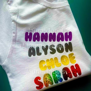 Puede incluir: Camiseta blanca con cuatro nombres impresos en letras coloridas y brillantes. Los nombres son Hannah, Alyson, Chloe y Sarah.