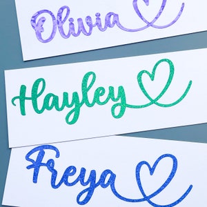 Pode incluir: Três nomes personalizados com corações de glitter. Os nomes são Olivia, Hayley e Freya. Os corações são de glitter roxo, verde e azul.