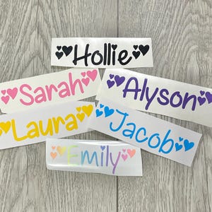 Puede incluir: Una colección de pegatinas de vinilo blanco con nombres en varios colores, incluyendo negro, rosa, amarillo, morado y azul. Cada nombre está acompañado de acentos en forma de corazón. Los nombres incluyen Hollie, Sarah, Laura, Alyson, Jacob y Emily.