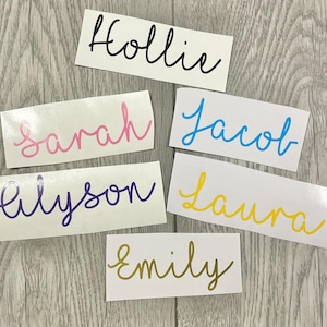 May include: A collection of white rectangular stickers, each bearing a name in a different colour. Names include Hollie, Sarah, Jacob, Alyson, Laura, and Emily, all written in a cursive script.