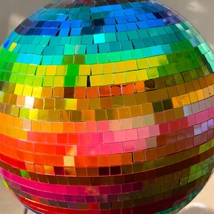 May include: A spherical disco ball covered in small, square, reflective mirror tiles arranged in horizontal bands of rainbow colors including red, orange, yellow, green, blue, and purple. A silver metal ring is attached to the top for hanging.