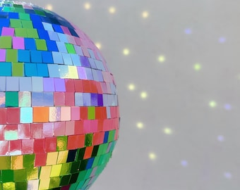 May include: A multi-colored disco ball with mirrored tiles hangs from a silver chain against a white background. The ball reflects light, creating a pattern of colorful dots on the wall.