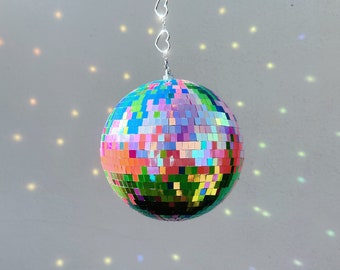May include: A multi-colored disco ball with mirrored tiles hangs from a silver chain with heart-shaped links. The ball casts rainbow-colored reflections on the white wall behind it.