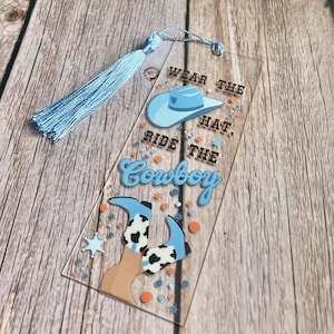 May include: A clear acrylic bookmark with a light blue tassel. The bookmark features a cowboy hat and boots design with the text "WEAR THE HAT. RIDE THE Cowboy". The design includes orange and blue accents.