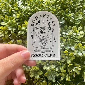 May include: White sticker with black text and illustrations. The text reads "ROMantasy BOOK CLUB". The illustrations include a fairy, a mermaid, bats, a crescent moon, a sword, and a book.
