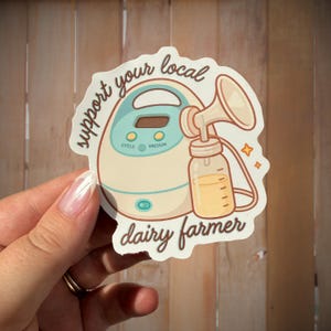 May include: A sticker with a cartoon breast pump illustration and the text "support your local dairy farmer." The pump is beige and teal, with a milk bottle. The sticker is held by a hand.