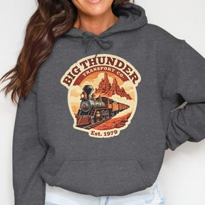 May include: Charcoal grey hoodie with a circular graphic depicting a vintage train, desert scenery, and the text "Big Thunder Transport Co. Est. 1979." The design uses orange, brown, and red.